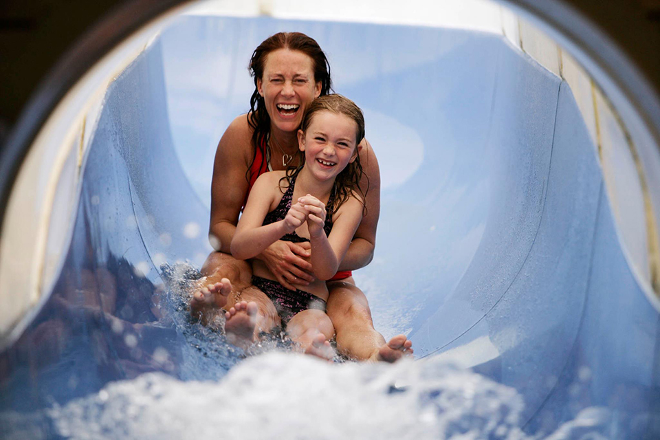 Bilden visar en kvinna och ett barn som åker ner tillsammans i en blå vattenrutschkana och skrattar. 
