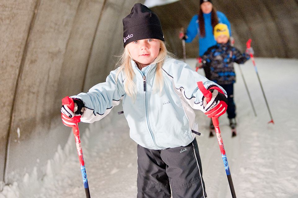 Bilden visar ett barn som åker längdskidor i skidtunneln. I bakgrunden åker ett till barn och en kvinna. 