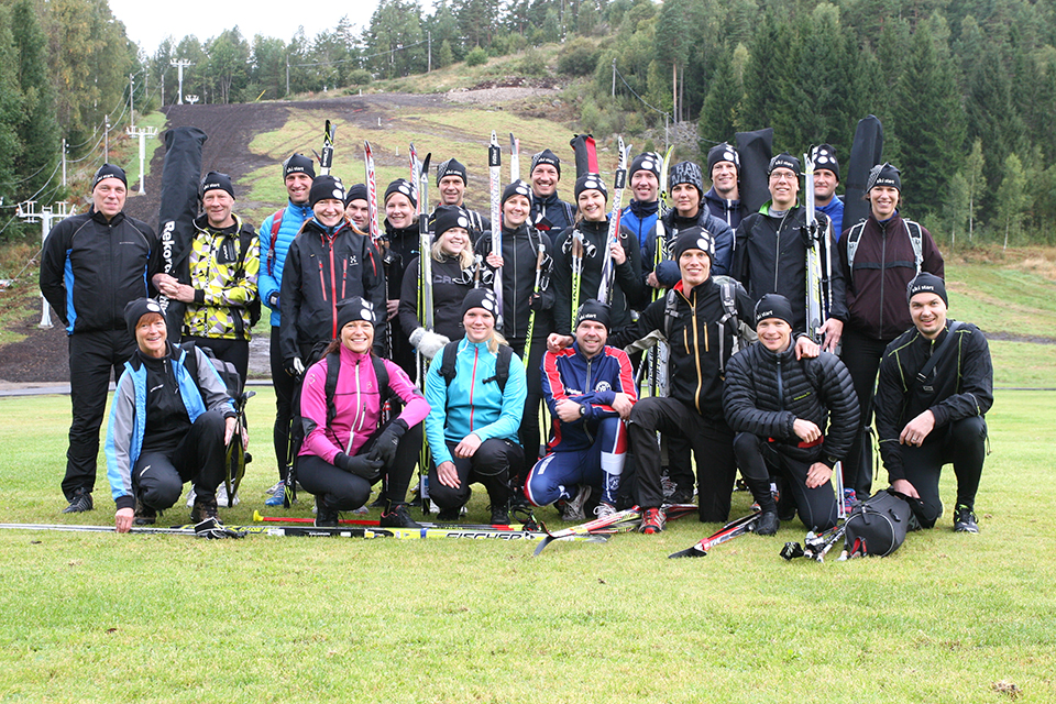Bilden visar en gruppbild på fotbollsplanen i Valberget under ett läger med Erik Wickström. Deltagarna står med skidorna bredvid sig och ler mot kameran. I bakgrunden ser man slalombacken. 